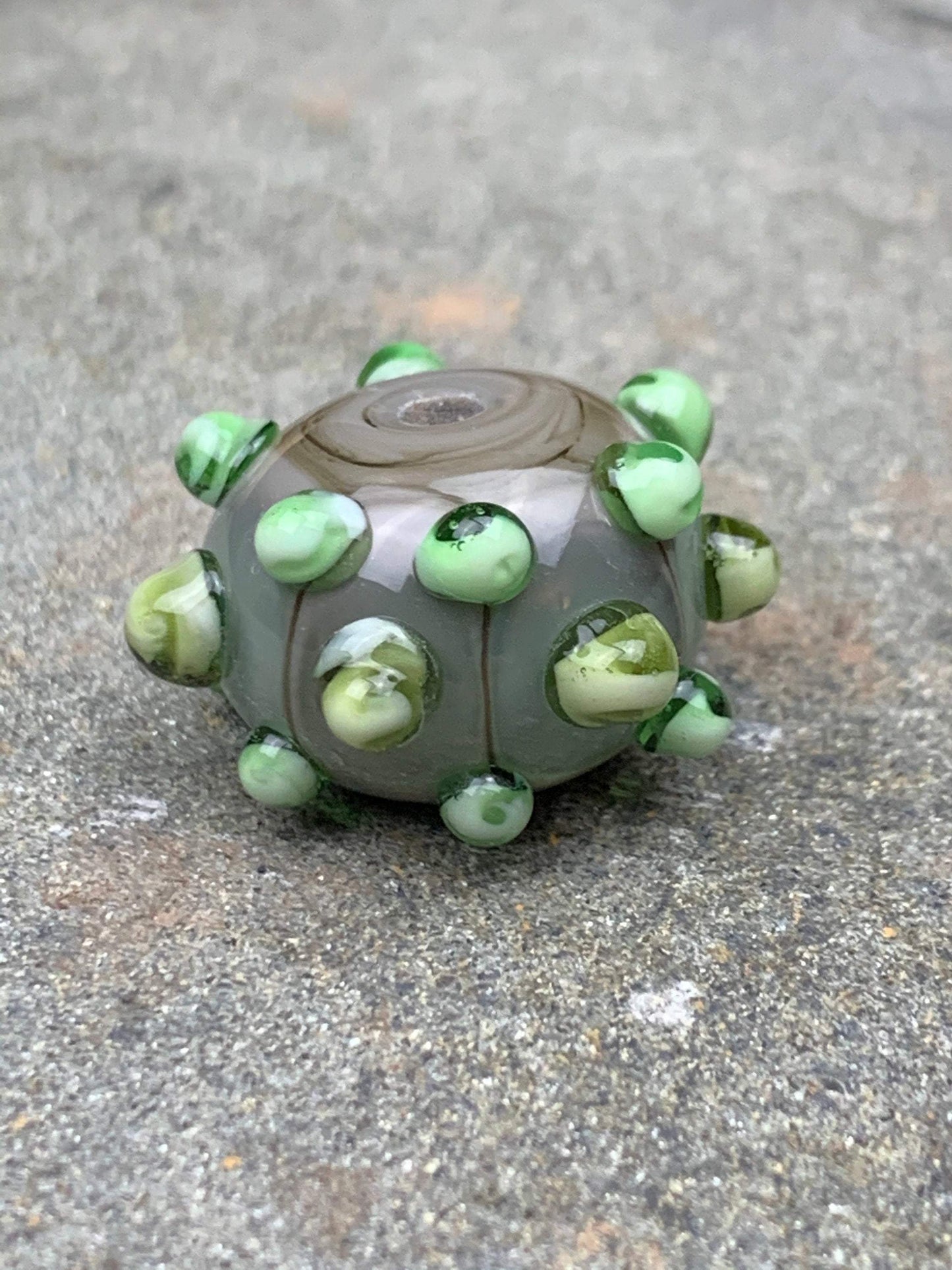 a handmade lampworked glass bead made of gray glass with green dots.