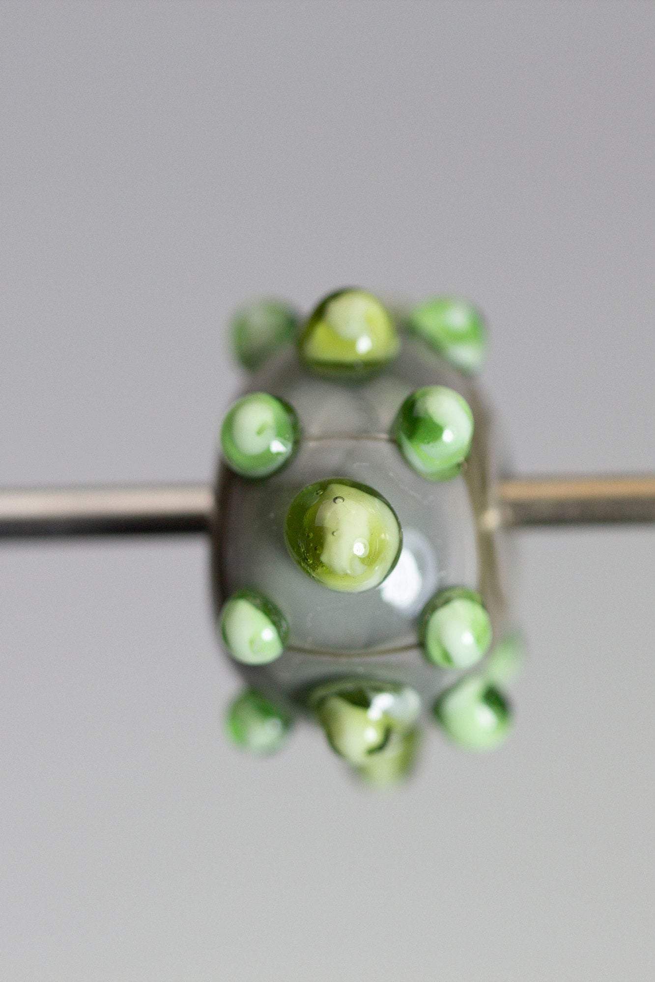 a handmade lampworked glass bead made of gray glass with green dots.