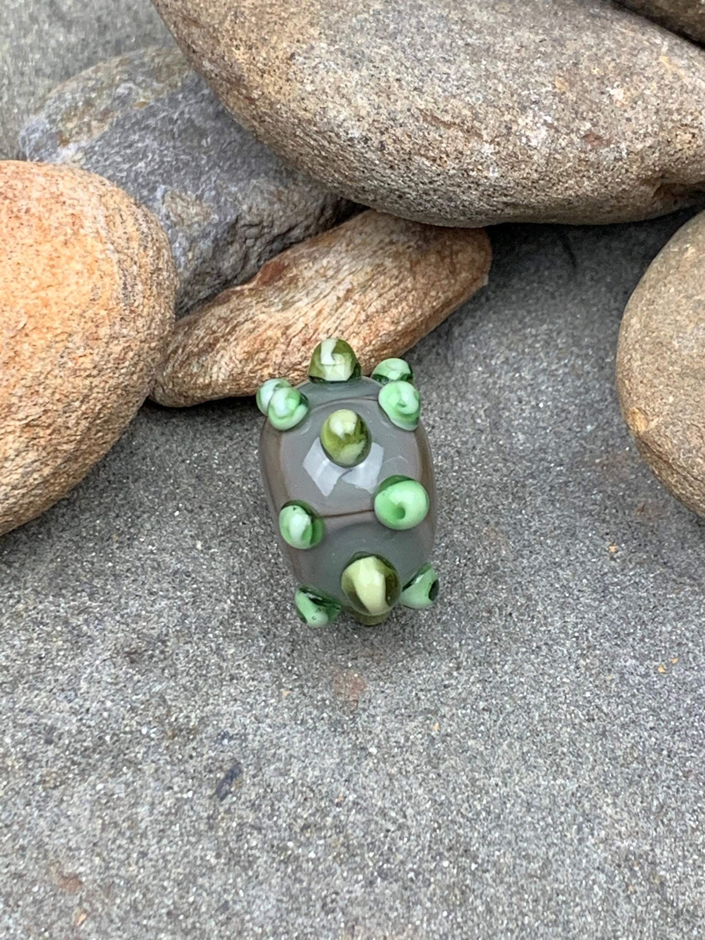 a handmade lampworked glass bead made of gray glass with green dots.