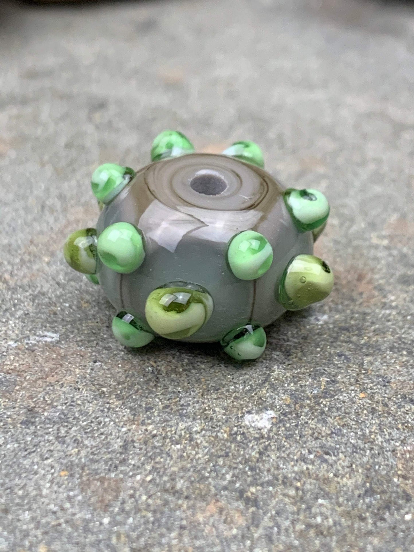 a handmade lampworked glass bead made of gray glass with green dots.