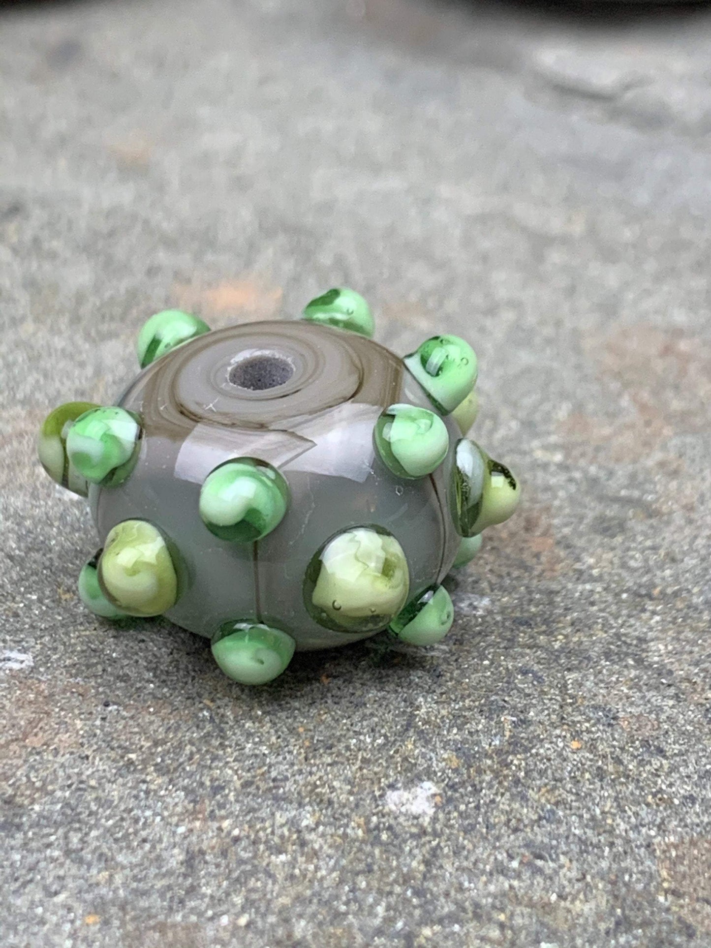 a handmade lampworked glass bead made of gray glass with green dots.