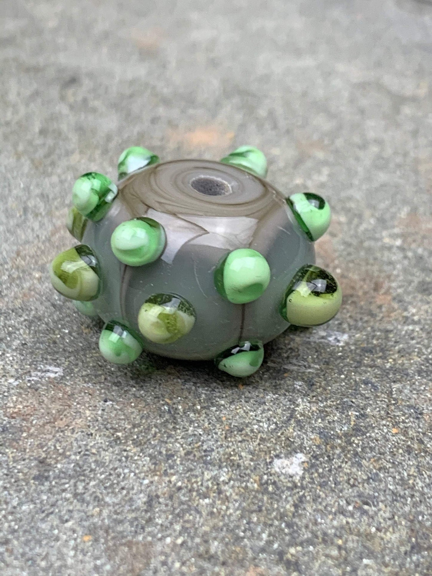 a handmade lampworked glass bead made of gray glass with green dots.
