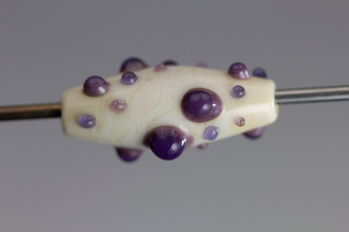 a handmade lampworked glass bead with purple and mauve dots on a white background