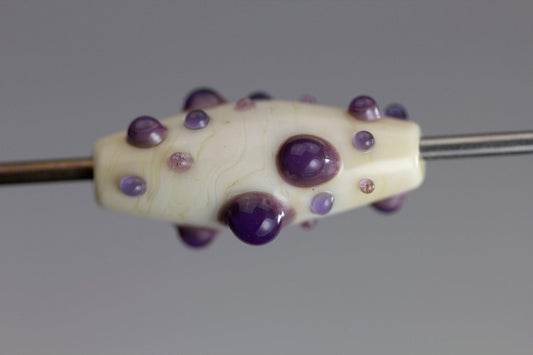 a handmade lampworked glass bead with purple and mauve dots on a white background