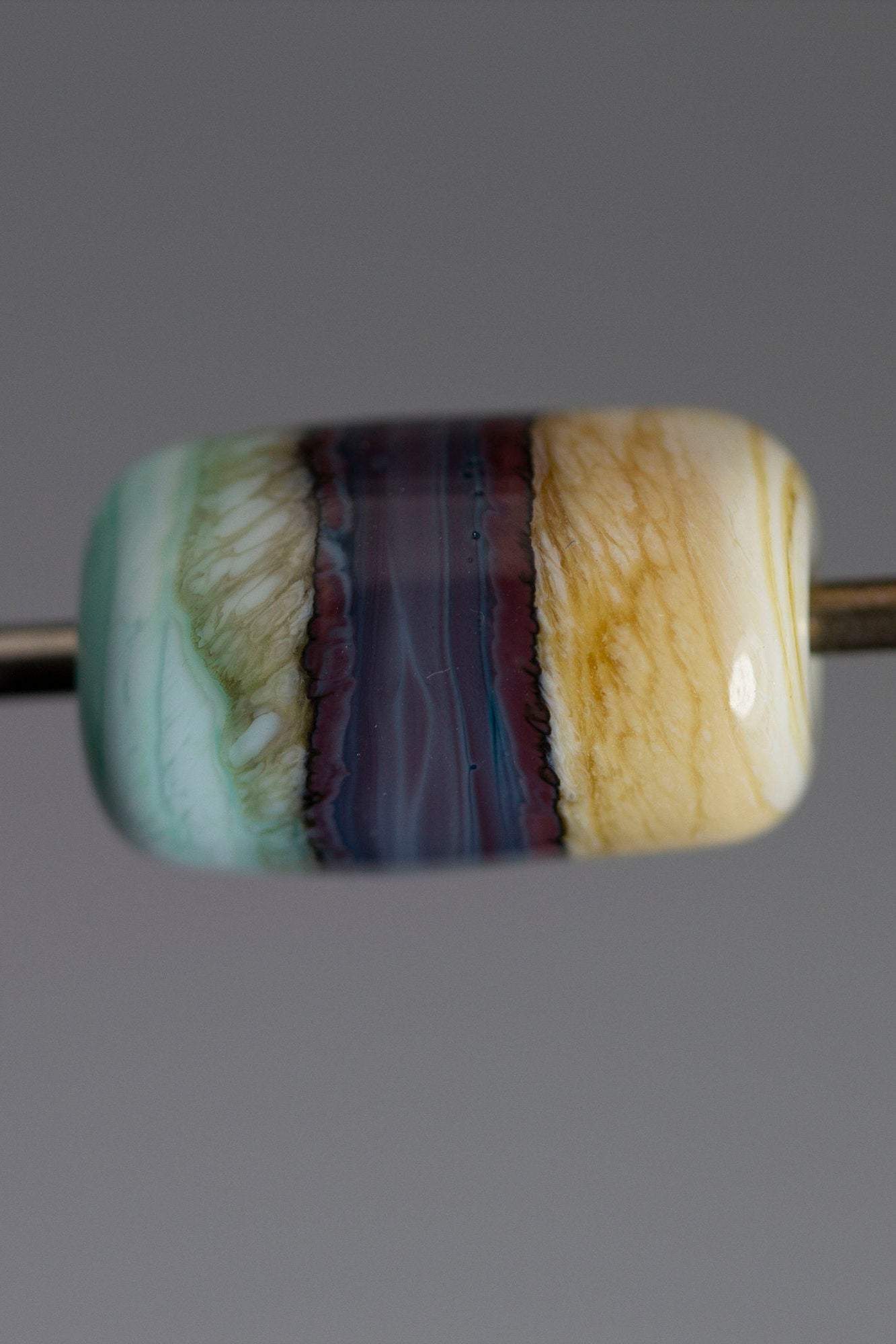 a handmade lampworked glass bead made of ivory, purple, and turquoise glass swirled into bands.