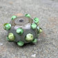 a handmade lampworked glass bead made of gray glass with green dots.