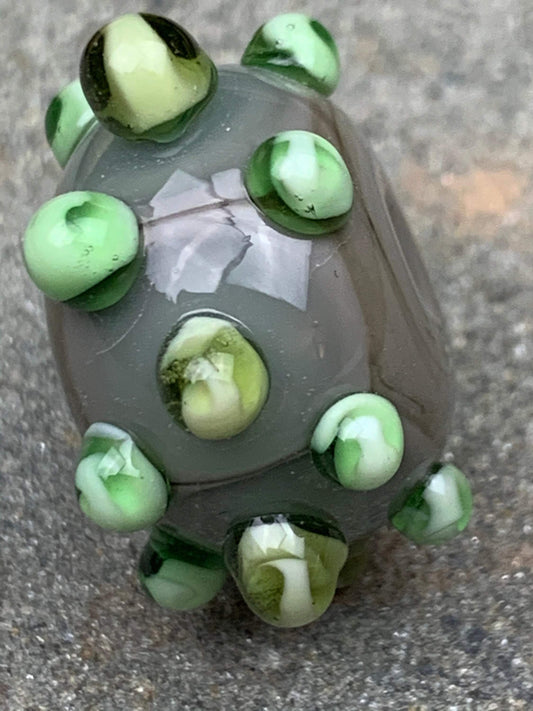 a handmade lampworked glass bead made of gray glass with green dots.