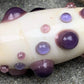 a handmade lampworked glass bead with purple and mauve dots on a white background